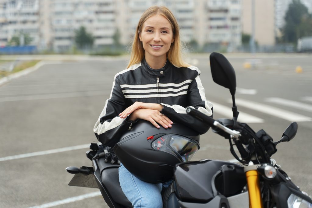 Woman in motorcycle gear smiling while sitting on a motorbike in an empty parking lot. Scooter Advice 360
