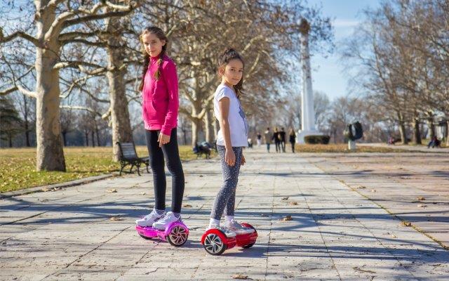 Two girls at a park, one riding a pink hoverboard and the other standing, with trees and a clear sky in the background. Scooter Advice 360