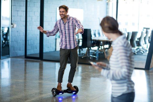 Man balancing on a hoverboard with open arms, smiling, as a woman looks at her phone in the foreground. Scooter Advice 360