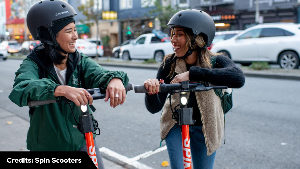 Two women are riding scooters on a city street, demonstrating how to use spin scooter. Scooter Advice