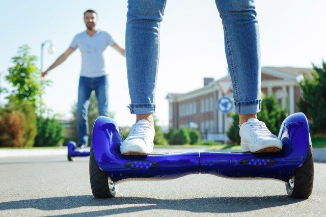 Person riding a blue hoverboard outdoors with another person standing in the background. Scooter Advice 360