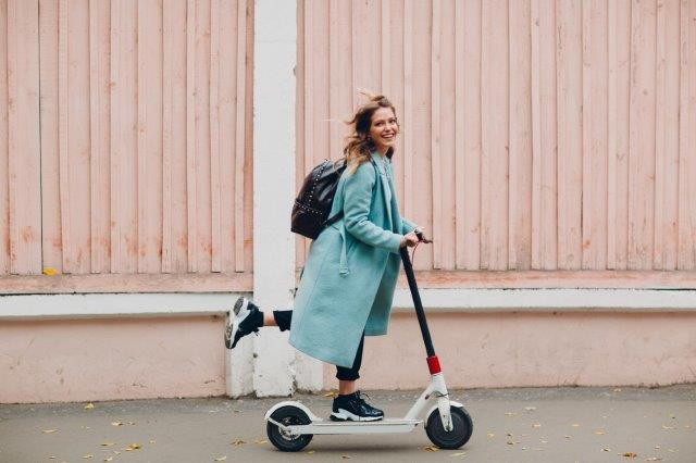 A woman riding an electric scooter in front of a pink wall. Are Electric Scooters Waterproof? Scooter Advice 360