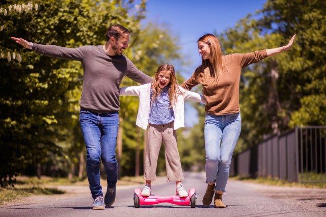 A child balancing on a hoverboard with arms outstretched, assisted by two smiling adults on a sunny day. Scooter Advice 360