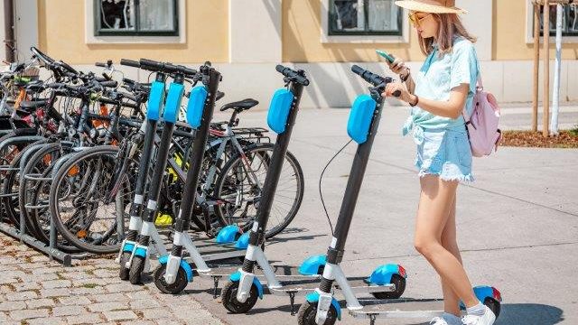 A woman looking at her phone while standing next to a group of electric scooters. Scooter Advice 360